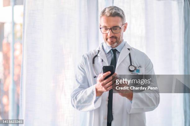 mature male doctor using a phone in a modern hospital room. one senior physician giving online medical advice on confidential communication chat app. healthcare worker using telemedicine application - doctor house call stock pictures, royalty-free photos & images