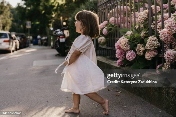 a cute toddler girl spinning barefoot in a white dress surrounded by hydrangea - kids fashion stock-fotos und bilder