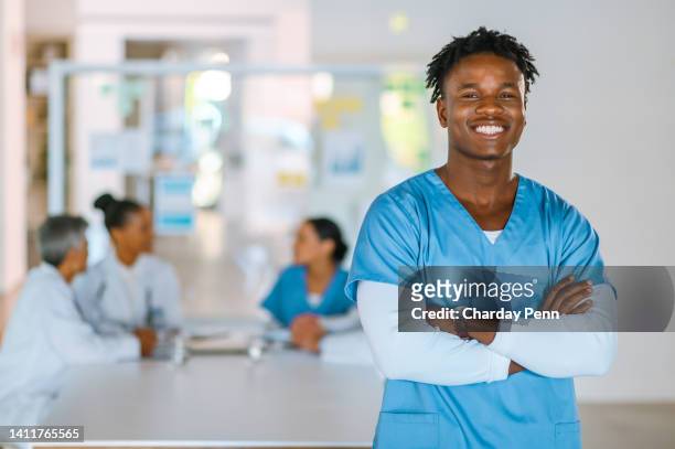 portrait of confident male doctor standing with folded arms and wearing scrubs at a medical meeting. proud african
healthcare worker or nurse smiling with colleagues working in the background - portrait background stockfoto's en -beelden