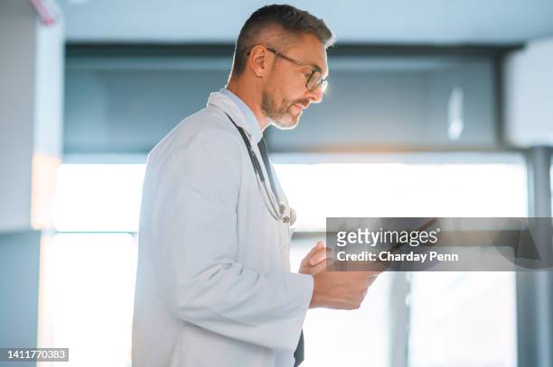 mature male doctor using a digital tablet for research online while working at a hospital alone. one serious healthcare worker searching the internet about an illness or disease to find a cure - doctor looking away stock pictures, royalty-free photos & images