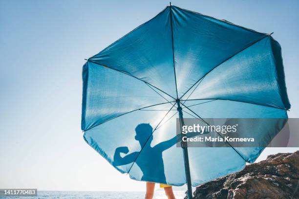silhouettes of boy flexing his muscles on blue beach umbrella. - summer refreshment stock pictures, royalty-free photos & images