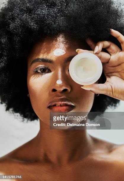 african female beauty model with an afro promoting lotion for skin care. beautiful black woman doing her skincare routine covering her eye with serum. portrait of a lady applying cream - applying imagens e fotografias de stock