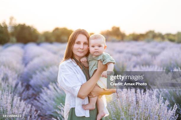 junge frau und ihre tochter im sommerkleid und sonnenhut gehen und umarmen sich bei sonnenuntergang durch das lavendelfeld - baby sun hat stock-fotos und bilder