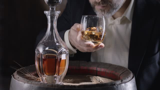 Man in a suit and beard (sommelier) takes a glass of whiskey on ice from a vintage whiskey barrel and shakes the glass to spread the smell of whiskey.