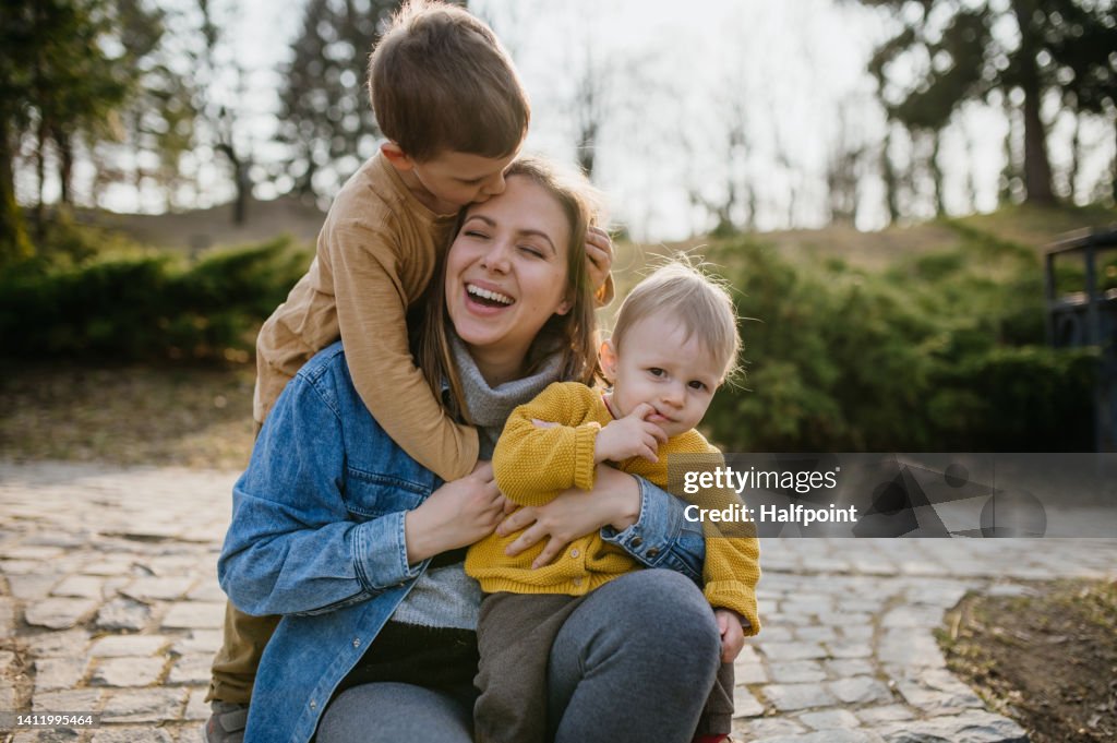 Happy young mother squatting and holding her little children, having fun, laughing in park in autumn.