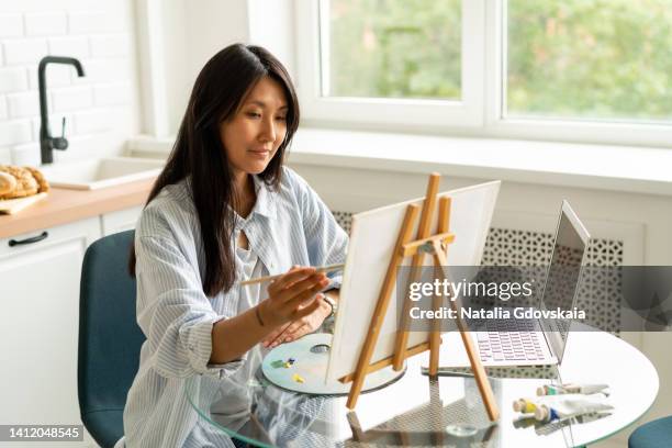 woman online learning painting skill. adult asian female oil drawing on easel in kitchen. following remote art course concept. improving knowledge at home. relaxing leisure activity. smiling artist creative occupation. holding paintbrush in hand - maler staffelei stock-fotos und bilder