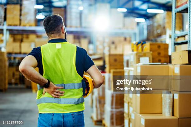 a warehouse worker having back pain and rubbing it. - ferido imagens e fotografias de stock