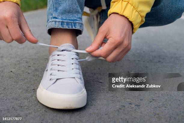 the girl's feet are shod in new sneakers and jeans. a woman in sports shoes squats down and ties or laces the untied lace. fashionable and stylish lifestyle. the concept of an active lifestyle, hiking. daily walking for health. - untied shoelace stock pictures, royalty-free photos & images