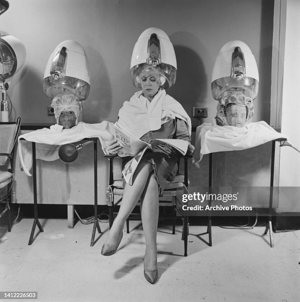 Woman, a towel draped over her shoulders, sits reading a magazine, her head under a rigid-hood hair dryer, while either side of her are two heads on...