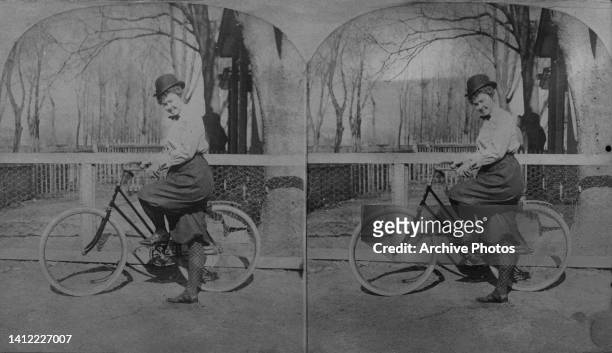 Stereoscopic image showing a woman, wearing plus fours and white blouse and a bowler hat, posing beside a bicycle, one foot on the pedal the other on...