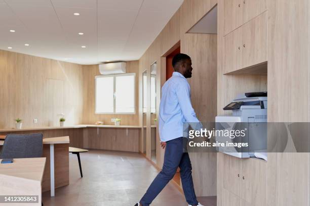 black man walking towards a door in a large office with a printer on the shelf with windows back - large printer stock pictures, royalty-free photos & images