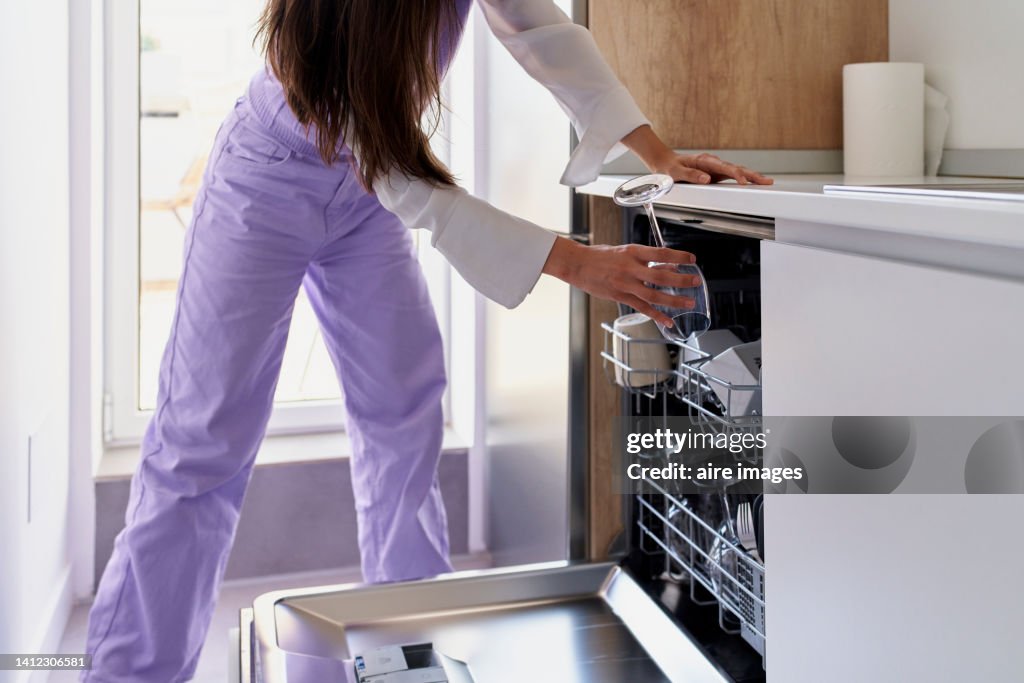 A white woman with long hair in purple pants and white blouse puts crockery to wash in the dishwasher