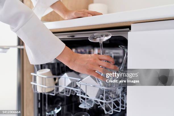 a white woman with long hair in purple pants and white blouse puts crockery to wash in the dishwasher - weinglas stock-fotos und bilder