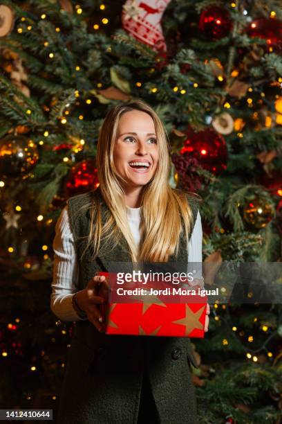 happy woman holding a christmas present wrapped in red wrapping paper laughing with a christmas tree behind her - giving tree stock pictures, royalty-free photos & images