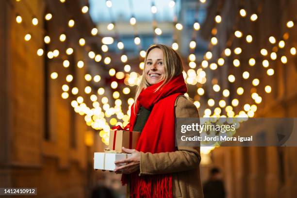 a happy standing woman holding christmas gifts with christmas lights in background - woman ribbon happy stockfoto's en -beelden