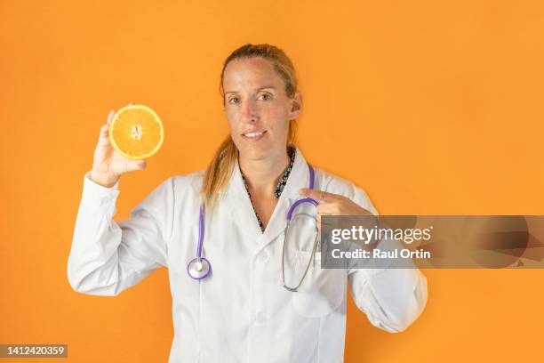 portrait of woman doctor wearing white coat and stethoscope holding and pointing to an orange fruit . - orange coat ストックフォトと画像