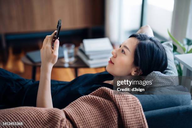 carefree young asian woman using smartphone while relaxing on sofa at cozy home. technology in everyday life. lifestyle and technology - 心地よい ストックフォトと画像