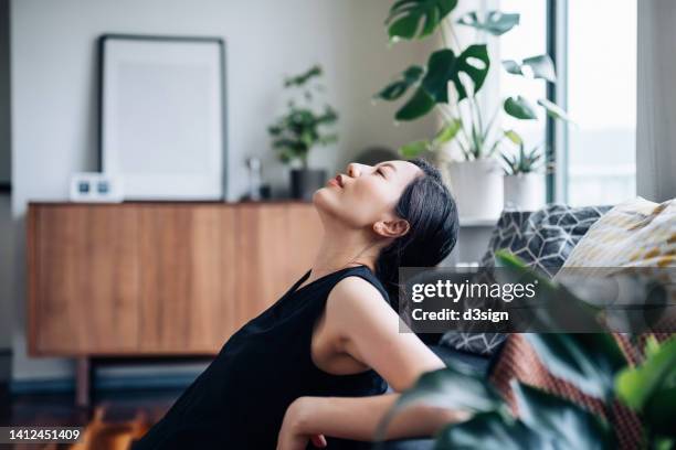 portrait of a beautiful young asian woman relaxes by the sofa in the living room, with her eyes closed and head up, enjoying the tranquility during leisure time at cozy home - gente tranquila fotografías e imágenes de stock