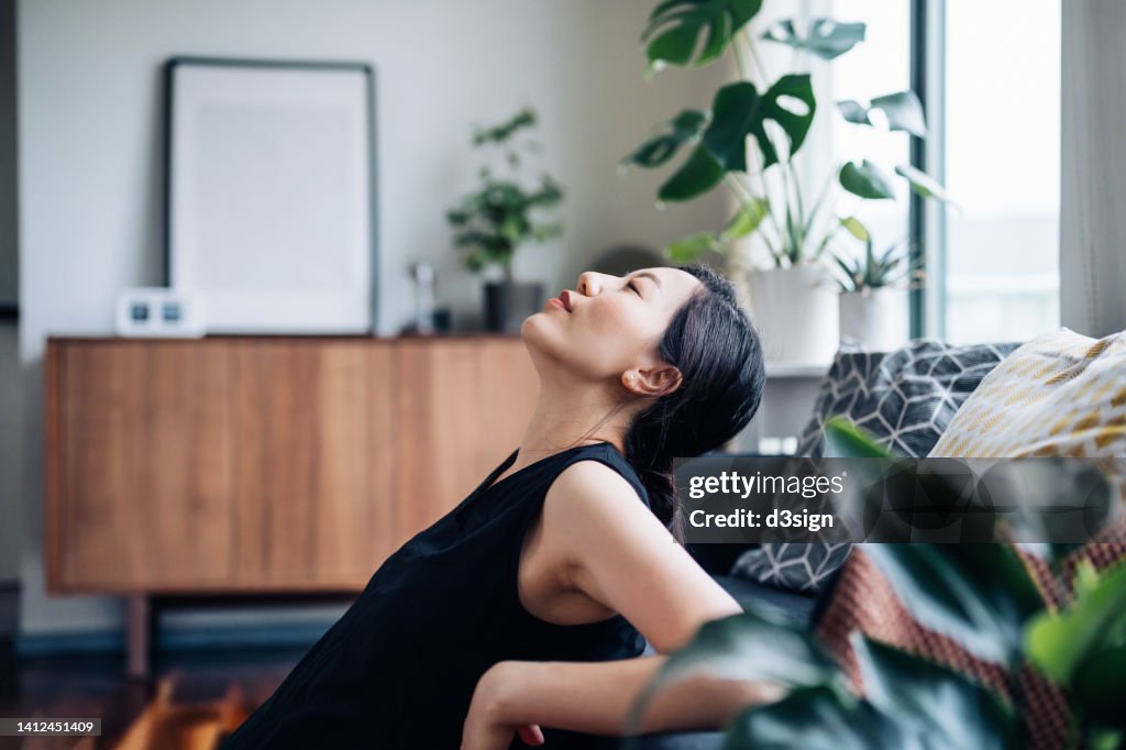 Portrait of a beautiful young Asian woman relaxes by the sofa in the living room, with her eyes closed and head up, enjoying the tranquility during leisure time at cozy home