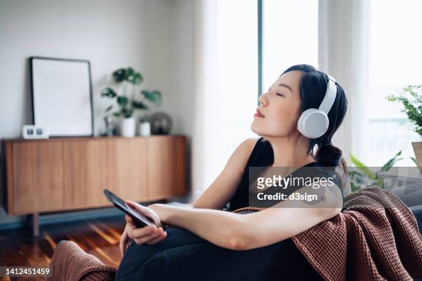 beautiful young asian woman with eyes closed enjoying music over headphones from her smartphone while relaxing on the sofa at cozy home - home sweet home stockfoto's en -beelden