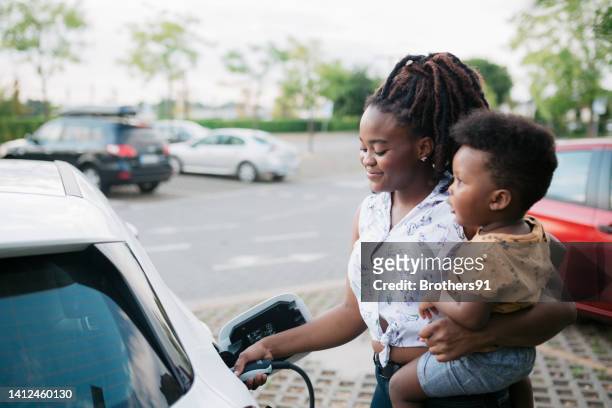 madre sosteniendo a su hijo mientras carga su vehículo eléctrico - cargar fotografías e imágenes de stock