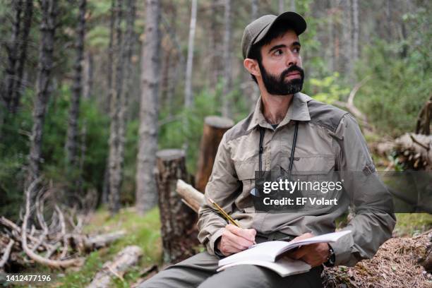 park ranger sitting in the forest writing notes on notebook looking to the side - schreibgerät stock-fotos und bilder