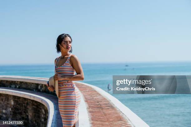 a young woman standing on the surface of a retaining wall looking back on a sunny day with sea on the background - guipuzcoa province stock pictures, royalty-free photos & images