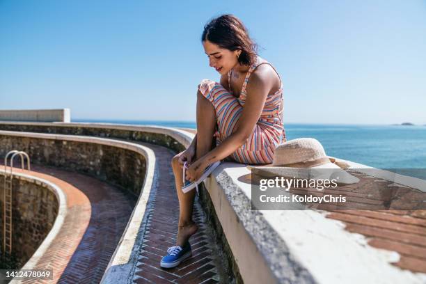 young woman tying her shoelaces sitting on retaining wall on the coast - shoelace fotografías e imágenes de stock