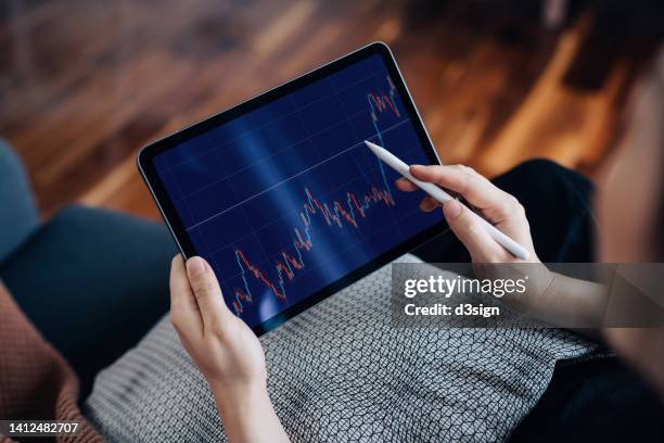 over the shoulder view of young asian woman managing finance and investment, analyzing stock market data on digital tablet at home. stock exchange, banking, finance, investment, financial trading concept. smart banking with technology - exchange rates stock pictures, royalty-free photos & images