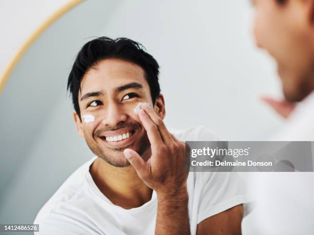 mann, der gesichtspflegecreme aufträgt, während er sich pflegt, schaut in seiner morgenroutine zu hause in den spiegel im badezimmer. nahaufnahme porträt eines gutaussehenden mannes mit gesichtslotion für saubere und klare haut - spf 30 stock-fotos und bilder