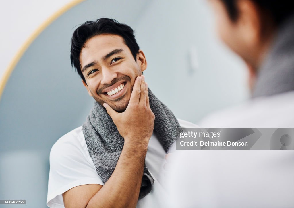 Grooming, hygiene and healthy facial skincare routine for smooth and soft male skin. Handsome young man looking in the mirror touching his beard happy about his facial shaving product