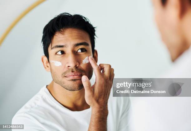 hombre que se aplica crema, loción y crema hidratante para una rutina de cuidado de la piel mientras se acicala en un espejo en casa. chico joven guapo que usa loción de protector solar con spf para protección uv en su cara para una piel sana - applying fotografías e imágenes de stock