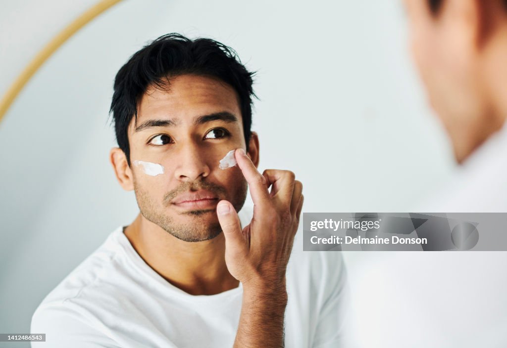 Man applying cream, lotion and moisturizer for a skincare routine while grooming in a mirror at home. Handsome young guy using sunscreen lotion with spf for uv protection on his face for healthy skin