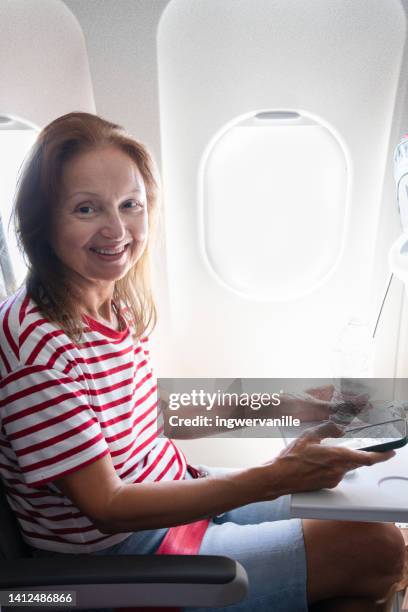 mature woman sitting by airplane window while using smart phone - thessaloniki airport stock pictures, royalty-free photos & images