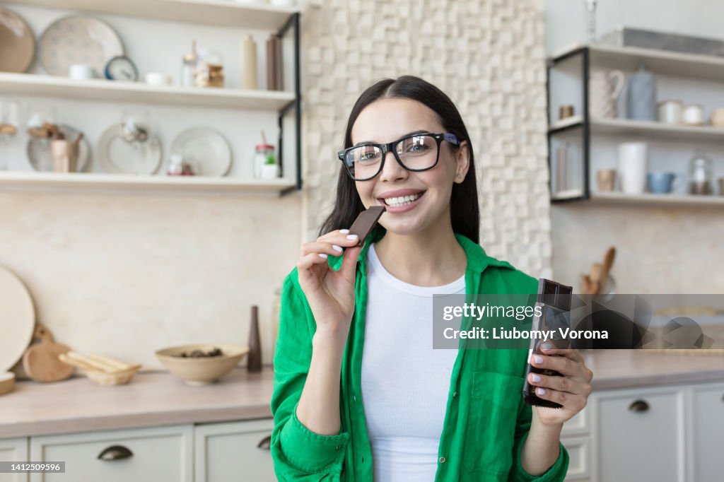 Young beautiful brunette woman in glasses and green shirt at home in kitchen, eating chocolate