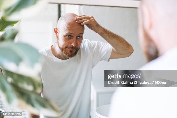 young adult bearded man looking in mirror in bathroom touching head worried about about hair loss - domestic bathroom - fotografias e filmes do acervo