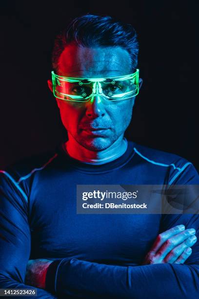 man with cybernetic glasses with green lights and a tight blue t-shirt, with his arms crossed, in a blue and red environment, and a black background. robot, cyborg, future and metaverse concept. - asimo stock pictures, royalty-free photos & images