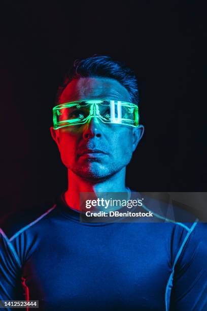 man with cybernetic glasses with green lights and a tight blue t-shirt, looking up, in a blue and red environment, and a black background. robot, cyborg, future and metaverse concept. - asimo stock pictures, royalty-free photos & images