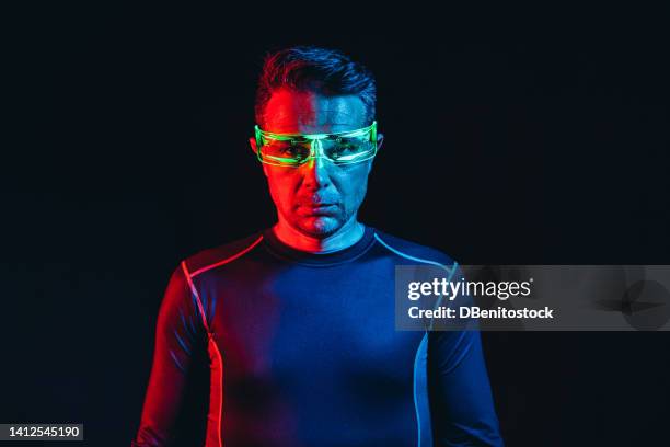 man with cybernetic glasses with green lights and a tight blue t-shirt, looking at the camera in a blue and red environment, and a black background. robot, cyborg, future and metaverse concept. - asimo stock pictures, royalty-free photos & images