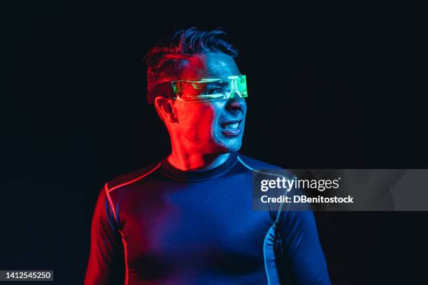 man with cyber glasses with green lights and tight blue t-shirt, screaming and looking to the side, in a blue and red environment, and black background. robot, cyborg, future and metaverse concept. - asimo stock pictures, royalty-free photos & images