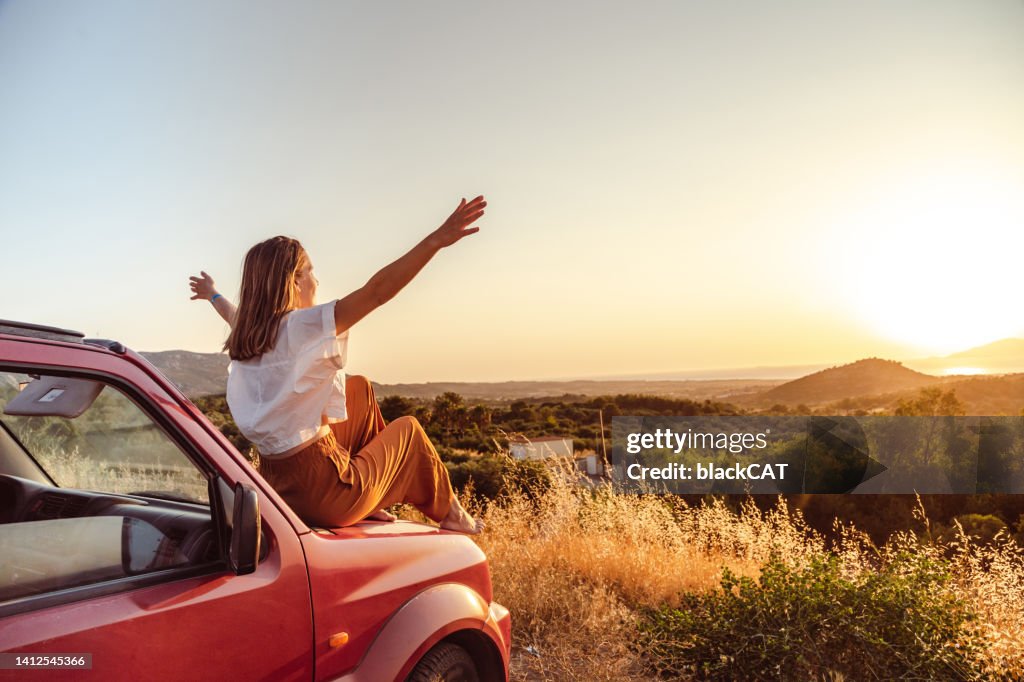 Joven con los brazos en alto sentada en el coche y disfrutando de la puesta de sol