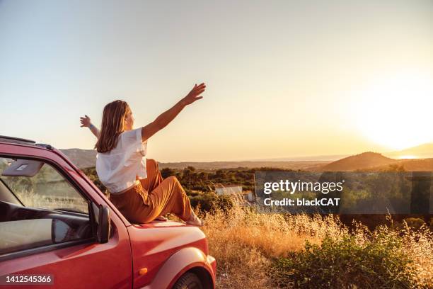 joven con los brazos en alto sentada en el coche y disfrutando de la puesta de sol - 4x4 fotografías e imágenes de stock