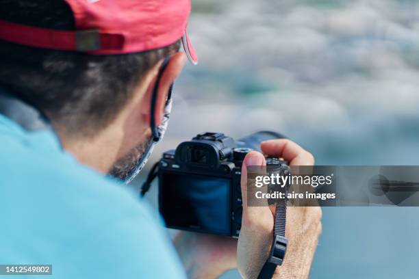 rear view of a tourist wearing mask walking and watching sunset looking away while holding camera on hill by sea against sky down at the beach - male photographer stock pictures, royalty-free photos & images