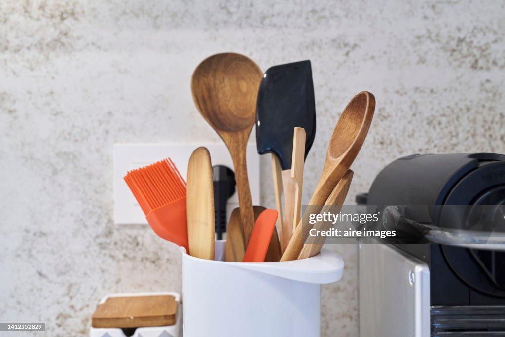 Close-up of front view of different kitchen utensils inside white container against a textured wall on worktop