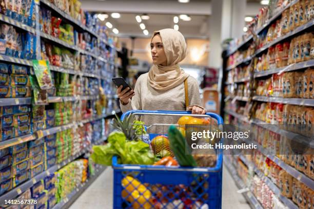 muslim woman buying groceries at the supermarket following a shopping list - shopping list trolley stock pictures, royalty-free photos & images