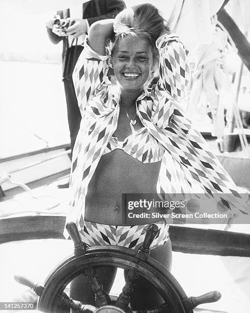 Jeanne Moreau, French actress, wearing a diamond print pattern bikini and matching blouse, smiling as she poses by the wheel of a boat, with her arms...