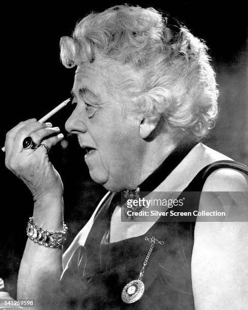 Margaret Rutherford , British actress, wearing a black choker around her neck, from which hangs a pendant, applying make-up to her eyes, in a...