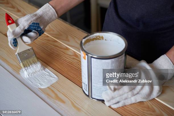 a brush with white paint in the hands of a woman or a man wearing protective gloves. painting and repair work. a female artist paints a board and a surface. a carpenter or builder paints boards. the concept of home or professional repair and construction. - white glove stock-fotos und bilder
