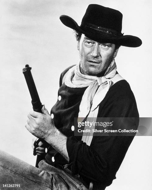 John Wayne , US actor, wearing a black cowboy hat and a white neckerchief, holding a rifle in a studio portrait, against a white background, issued...