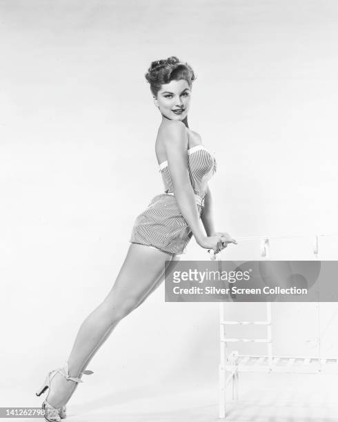 Full length portrait of Debra Paget, US actress, wearing a striped swimsuit, leaning against the back rest of a chair in a studio portrait, against a...